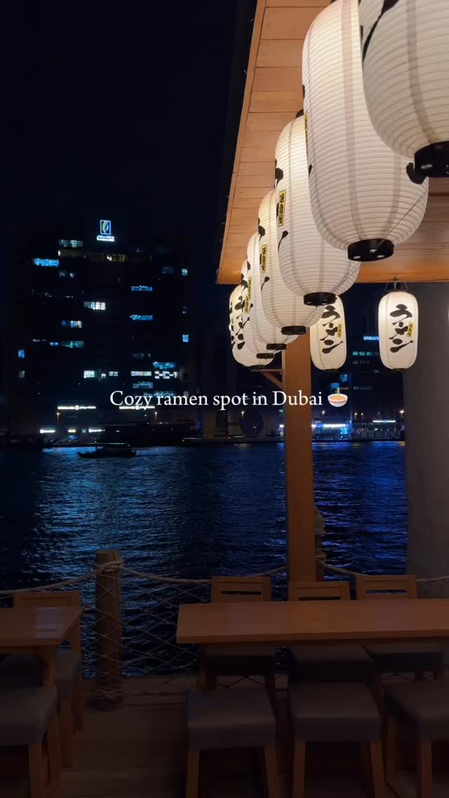 A nighttime scene at a ramen restaurant in Dubai. The image is dominated by dark blues and blacks, with the water of a body of water prominent in the middle ground. Overhead, a line of Japanese paper lanterns hang, casting a warm glow on the scene. In the foreground, wooden tables and chairs are visible, implying an outdoor seating area. The overall impression is one of a cozy, inviting restaurant with beautiful views.