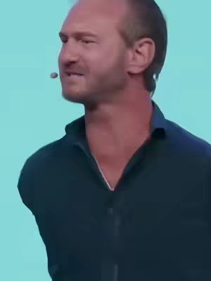 The image features a man speaking with a microphone clipped to his shirt. The background is a solid light-blue. He is slightly off-center in the frame with his face angled towards the left.