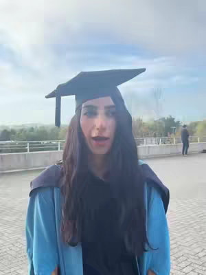 A woman in a graduation cap and gown stands outdoors, looking directly at the camera with a slightly open mouth. The background shows a blurred landscape under a cloudy sky. The woman's attire includes a black graduation cap and a light blue gown.