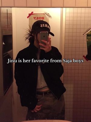 A person is taking a selfie in a bathroom mirror. The person is wearing a black cap, black jacket, a white sleeveless shirt, and brown pants. A text overlay states, "Jinu is her favorite from Saja boys."