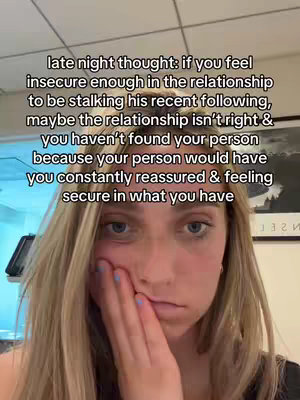 A selfie-style image featuring a young woman with blonde hair, looking at the camera with a slightly pensive expression. She has her left hand, with blue nail polish, delicately touching her cheek. The background is blurred, but appears to be an indoor setting.
