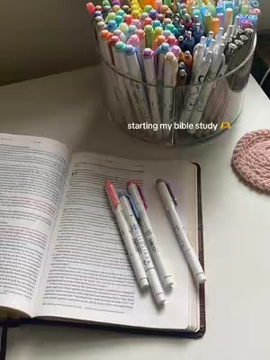 An open Bible with colorful pens lying on top and a container full of similar pens in near proximity. The shot is well-lit presenting a sense of calm.