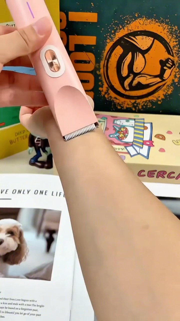 A close-up shot of a person's arm with a pink electric razor in use. The razor is shaving a small area of the arm. The background is blurred, showing items on a desk, including a book, a box, and a logo.