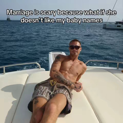 An image of a man, shirtless and wearing sunglasses, lounging on what appears to be the deck of a yacht. He is reclining on a white surface with a blue ocean and a few boats in the background.