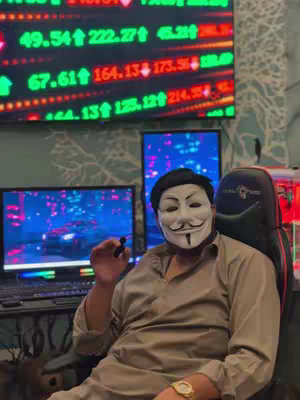 The cover image shows a person wearing a Guy Fawkes mask, sitting in what appears to be a gaming chair in front of multiple computer screens. Behind them, there's a large screen displaying what looks to be financial data, with green and red numbers and a backdrop of a patterned wall. The individual is holding a small item, possibly a pen, with one arm resting on the armrest of the chair. There are also screens showing a car on a road.