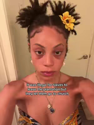 A selfie-style shot of a young woman with dark skin and curly hair styled in two buns, adorned with a yellow flower. She is looking directly at the camera with a neutral expression, wearing a necklace and a patterned top against a plain wall background. The text overlay presents a historical statement with a critical nuance.