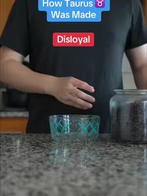 The cover image shows a person from the torso down at a counter surface. In the foreground, there is a clear, patterned glass bowl and to the right, a large glass jar partially filled with dark beans is visible. The person's hand is positioned above the bowl, and they are wearing a black shirt. The text overlay reads "How Taurus ♉ Was Made Disloyal". The color palette is neutral with varying shades of gray on the counter and a dark background.