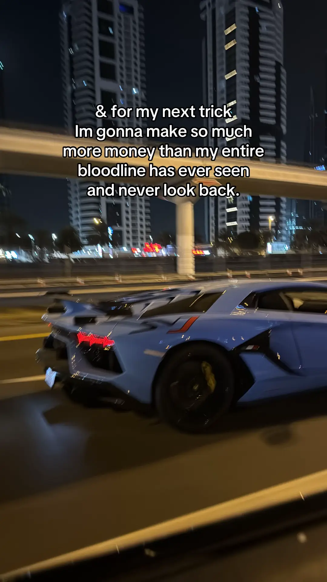 A night scene featuring a light blue Lamborghini Aventador driving on a road. The car is in motion, with its rear lights illuminated and visible. A tall, modern cityscape with illuminated buildings fills the background, suggesting an urban environment. The overall composition emphasizes speed and luxury.