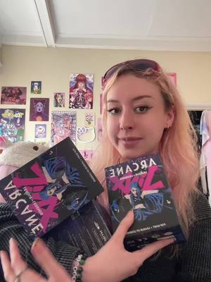 A young woman with pink hair is holding two boxes with 'Arcane' branding, looking at the camera with a smile. She is wearing glasses, and there are posters on the wall behind her. The photo is taken in a well-lit room with a casual atmosphere.