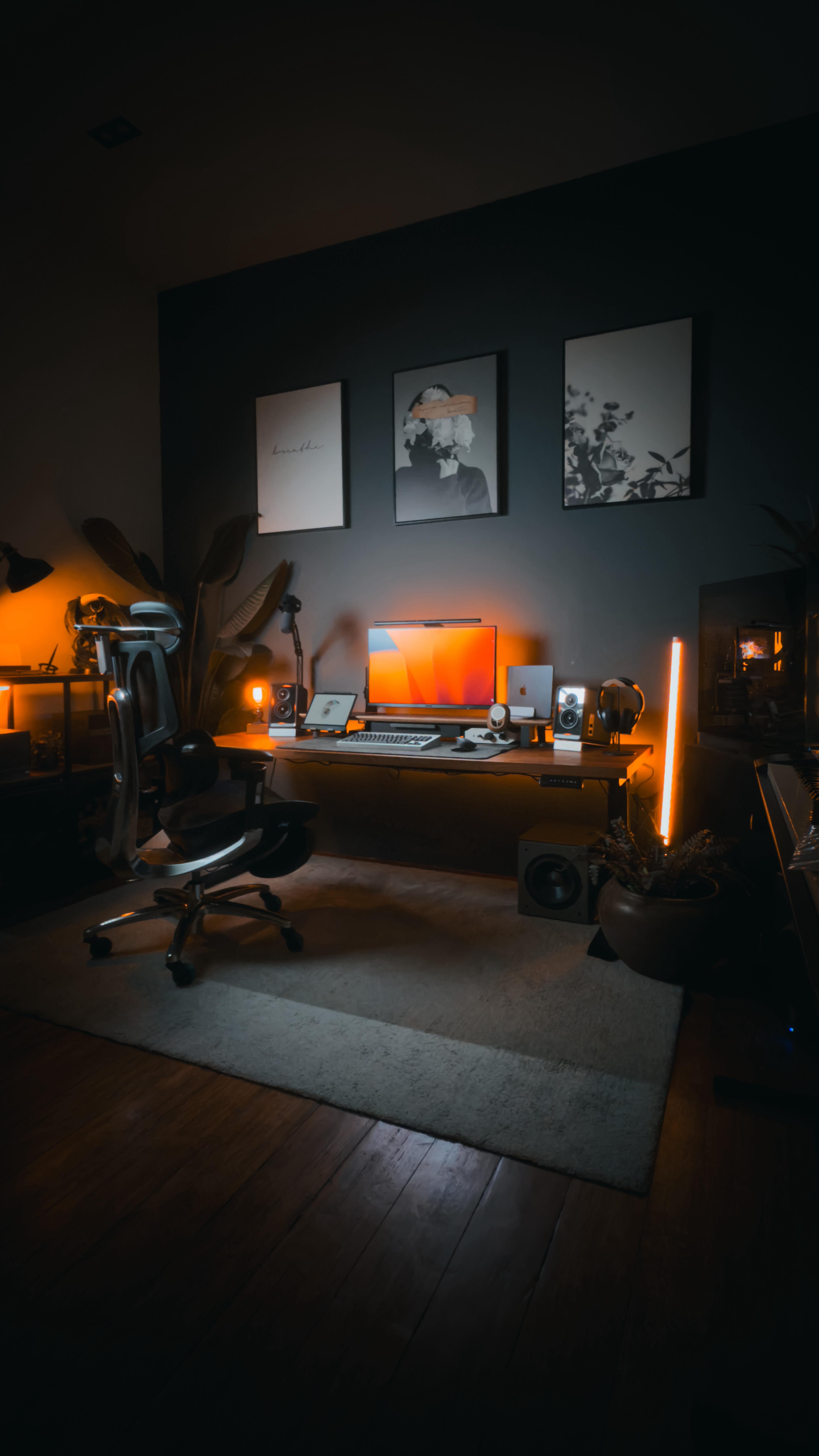 A dimly lit room with an organized workspace featuring a wooden desk. On the desk, there's a computer monitor with a vibrant orange display, a laptop, speakers, and various tech accessories. The room has dark walls adorned with framed artwork, decorative plants, and warm orange lighting from multiple lamps and a vertical light bar. A white rug covers part of the floor, adding contrast to the dark wooden flooring and walls.