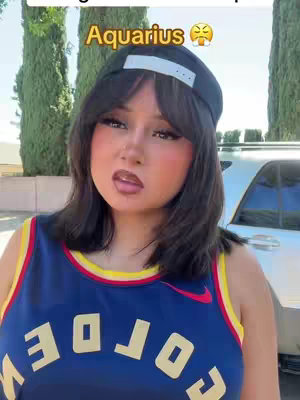 A woman with dark hair wearing a baseball cap and a basketball jersey is looking at the camera with an expression of discontent. Text in a gold box above her head says "Aquarius" with an emoji that appears angry.