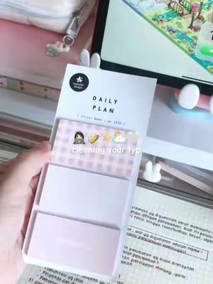 The image showcases a person's hand holding a white notepad featuring a daily plan template, emojis, and the text 'cleaning your typ'. The background includes a study setup with a laptop, a pen case, and decorative items like bunny ears. The overall aesthetic appears organized and study-focused, with a soft color palette.