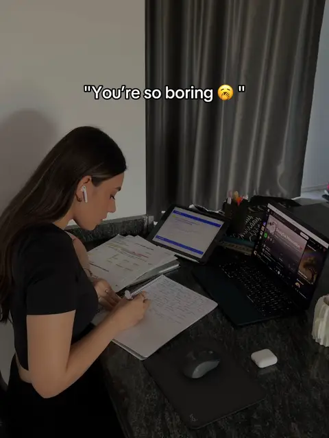 A woman is seated at a desk, writing in a notebook with a laptop and tablet open in front of her. Text overlays the image, and the room has neutral colors and a dark curtain.