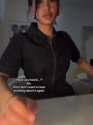A person, likely female, wearing a black collared shirt with a zipper, is positioned in the foreground. The image is lit with an overall dimness, and the background includes a faint glimpse of other objects, suggesting a home or indoor setting. The person's facial expression is slightly obscured, but the overall pose and tone suggest a conversation or reaction.