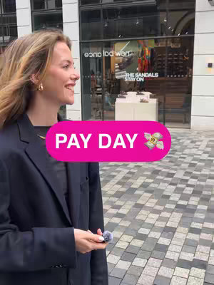 A woman is walking on a brick-paved street smiling towards the left, with a building and a shop entrance in the background. There is a pink banner with the text "PAY DAY" and a money bag icon placed in the center.