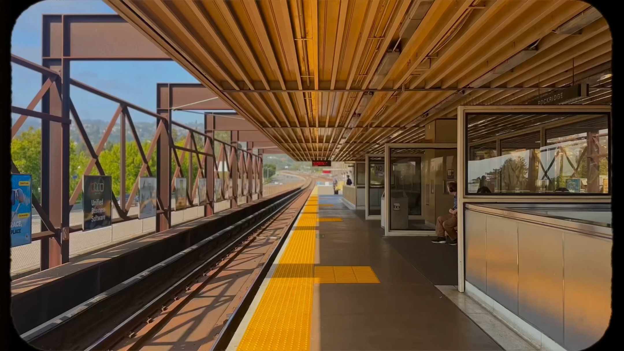 The cover image depicts a train platform. The foreground is dominated by yellow tactile paving and train tracks, with the perspective leading to the horizon. A steel overhead structure provides shelter where people can be seen waiting and a sign that says "Rockridge." The background shows a blue sky and the outline of hills.