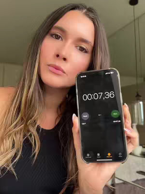 A woman with long, wavy hair is holding a phone displaying a timer. She is looking at the camera with a neutral expression. The background appears to be a white wall, and there are glimpses of household objects.