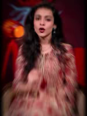 A blurred image of a woman with long, dark hair and a focused expression, possibly speaking or singing. She is wearing a patterned top, and the background is a deep red, with faint blurred shapes suggesting a setting or props.