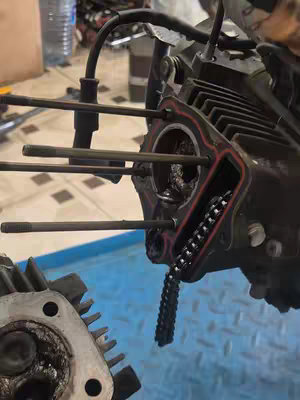An overhead shot showcases a disassembled engine, with one part detached and positioned to the left. The main component, a dark-colored engine block, has internal components exposed, including cylinder head studs and a chain. Other parts, like what appears to be the cylinder head, are placed nearby. The setting looks to be a workshop or garage with a blue surface below the engine.
