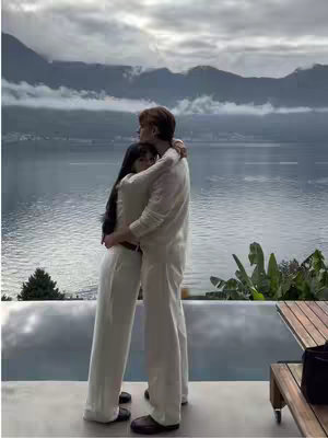 A couple embraces, seen from behind, overlooking a scenic landscape of mountains and water under a cloudy sky. The man wears a white outfit, and the woman, with long, dark hair, is also in white. They stand on a patio with a wooden table, plants, and the edge of what looks like a pool.
