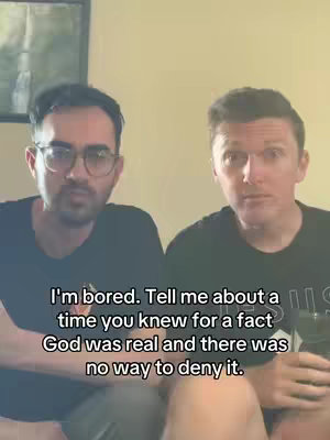 Two men in the image, sitting close, staring forward. The background is a plain wall. The image is well-lit, with a conversational feel.