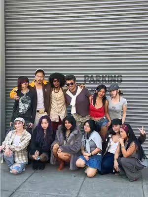 A group of about 10 people of diverse ages and clothing styles standing in front of a gray, horizontally-ridged, garage-style door. The group is informally posed, with some people in the front row crouching or seated. The clothing styles are eclectic, varying from casual to slightly more dressed up, with an emphasis on vintage or secondhand pieces. The color palette is varied, with pops of color amidst more neutral tones of the background.