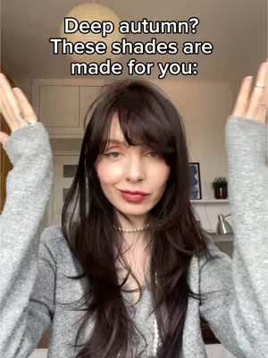 A woman with long, dark hair and a gray sweater stands in front of a neutral-toned background, arms raised and hands open. Text in white overlayed at the top reads "Deep autumn? These shades are made for you:"