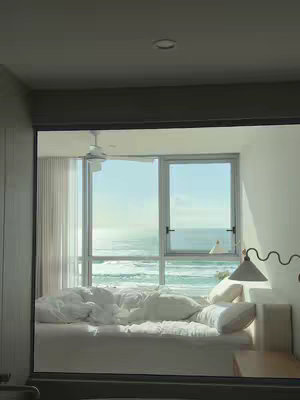 A serene scene of a bedroom with a large window overlooking the ocean. The bed is unmade, with white linens, suggesting a relaxed atmosphere. The sunlight streams in, illuminating the space and highlighting the view of the sea.