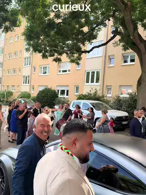 The cover image shows a candid shot of a wedding scene. In the foreground, a man is smiling and standing near a silver car. Another man, wearing a beige coat and a colorful lei, is walking in front of the car. Behind them, there are people gathered and dressed in various outfits, suggesting a social gathering or event. Buildings and some trees form the backdrop, creating an outdoor setting.