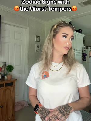 A woman with blonde hair and sunglasses on top of her head is standing in a room. She wears a white t-shirt with a sunset graphic. The background includes a door, side table with a plant an art prints. Text on the cover image reads "Zodiac Signs with the Worst Tempers".