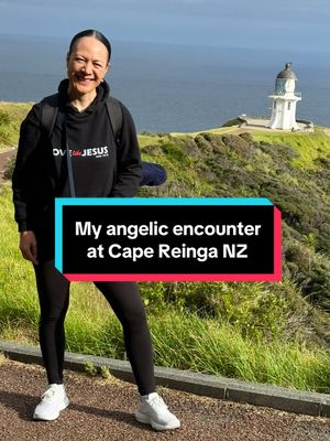 A woman stands on a path near a coastal landscape. She is center-frame, smiling at the viewer. Text overlayed on the image says 'My angelic encounter at Cape Reinga NZ'. Behind her, the ocean meets a rocky coastline, and a white lighthouse is perched atop the cliffs. The sky is overcast.