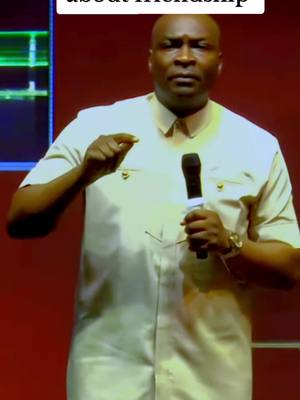 A man in a light-colored button-up shirt holding a microphone and gesturing with his right hand. He is standing in front of a red backdrop, and a microphone is visible. The image has a medium shot with even and balanced lighting.