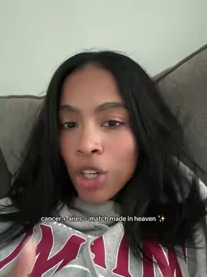 A woman with long, dark hair is centered in the frame, speaking directly to the camera. The background is a plain, light-colored surface. A text overlay appears at the bottom, and the woman is wearing a gray top.