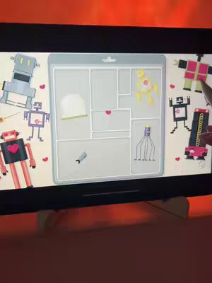 The cover image showcases a screen displaying the puzzle game "A Little to the Left." The screen is a gray tray containing various shaped compartments holding objects. Surrounding the tray are several cartoon robot illustrations. The background appears to be a warm orange.
