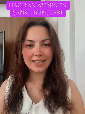 A woman with long brown hair is centered in the frame, her face lit brightly. She wears a white top. The background is interior, with parts of a door frame and wall visible. The image has a natural, well-lit quality.