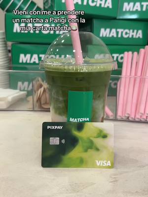 The image showcases a matcha latte inside a clear plastic cup, a pink straw is inserted. In the foreground is a credit card with the text "PIXPAY" and "VISA", near a green holder with the word "MATCHA". Behind the cup is a blurry background with more matcha and matcha packaging.