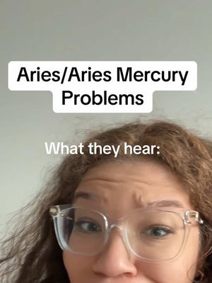 A selfie-style video cover image features a person, likely female, with curly brown hair and wearing clear-framed glasses. The top half of the frame displays text overlays. One white rectangular text box reads 'Aries/Aries Mercury Problems'. Below this, the text overlay 'What they hear:' appears in white text.