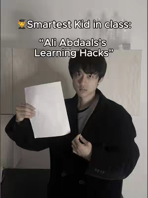 A young Asian man stands facing the camera, holding a blank white paper in front of him. He is dressed in a dark overcoat over a gray shirt. Above his head, white text overlay reads, "Smartest Kid in class:". Beneath this, centered text states "Ali Abdaals's Learning Hacks". The background is a plain, lightly colored wall, with a few objects on a shelf.