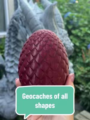 A person's hand holding a dark red, egg-shaped container in front of a blurred-out statue of a dragon head and greenery. A light green speech bubble with white text reads 'Geocaches of all shapes'.
