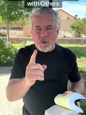 A middle-aged man with gray hair and a beard is outdoors, pointing his right index finger towards the camera. He is wearing a black t-shirt against a background of greenery and a building. The image is well-lit, suggesting daytime, and the man is looking directly at the camera. In the top bar, the text 'with Others' appears.