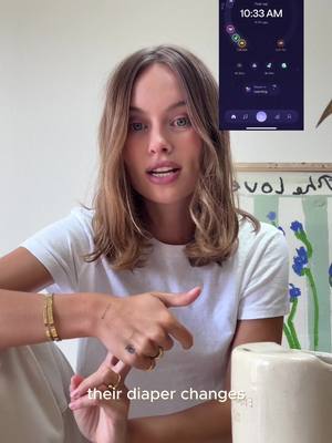 A young woman is centered in a well-lit room, looking directly at the camera and speaking. Her light brown hair is styled with loose waves. The image includes a cropped view of an app interface on a vertical smartphone, displaying what seems to be a time and some related functions. The woman is wearing a white t-shirt and a gold bracelet on her left wrist. Her right arm is bent and her finger is pointing downward, also wearing a ring and displaying a tattoo on her left hand. Behind her, a blurred painting or decoration with floral elements adds a touch of softness to the background.