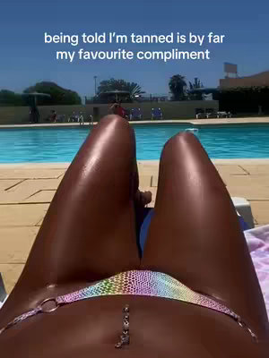 A first-person view of a person lying down, displaying their tanned legs from the waist down, wearing a colorful bikini, with a piercig and a shimmering light, positioned against a bright blue pool. In the background, people are sitting and trees are visible.