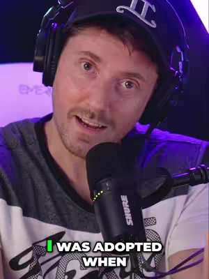 A man with brown hair wearing a black cap and headphones looks at the camera while speaking into a microphone. He's wearing a white and gray striped shirt and a text overlay says "I WAS ADOPTED WHEN".
