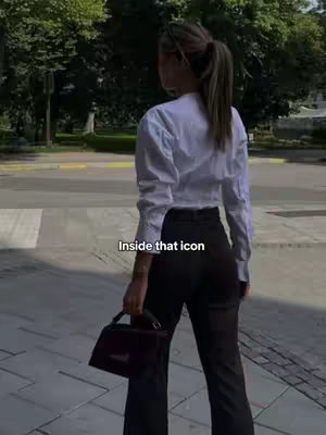 A woman with long hair in a ponytail is captured from behind, walking on a city street. She wears a white button-up shirt and dark pants, holding a purse, with trees and buildings in the background.