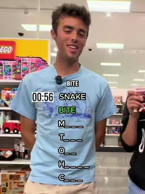A young man wearing a light blue t-shirt stands in what appears to be a retail store. A humorous interactive word puzzle is overlaid on his shirt. In the background, shelves are filled with colorful products and shopping items The overall lighting is bright, and the composition is vertically oriented.