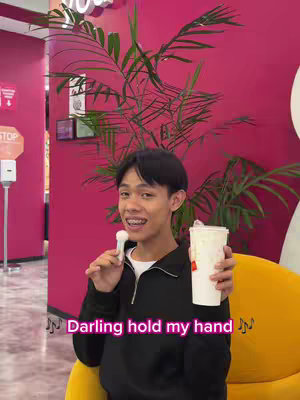 A young man with braces, wearing a black zip-up, is holding a small handheld microphone or candy in his left hand and a large beverage in his right hand. He is positioned in front of a pink wall with green plants as a background, with a yellow chair visible.