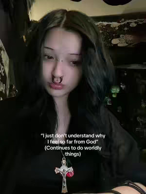 A close-up selfie of a person with dark hair, multiple piercings, and a cross necklace. The person is looking down with a slightly sad expression. Text overlayed on the image expresses a feeling of distance from God.