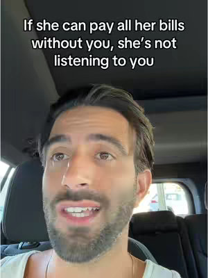 A selfie-style video shows a man speaking while seated in a car. The text overlay at the top of the image reads: 'If she can pay all her bills without you, she's not listening to you.' The man has short, dark hair, a trimmed beard, and is wearing a white shirt. The car's interior and a sliver of the sky are visible in the background.