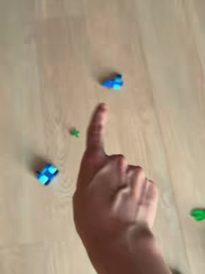 A first-person perspective shot with the creator's hand pointing down towards Lego pieces scattered on a wooden floor. Several blue and green Lego pieces are visible with the index finger pointing down, with a blur of motion.