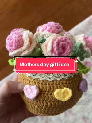 The image shows a close-up of a handmade crochet flower pot filled with crochet flowers. The pot is a dark brown color with heart-shaped embellishments. The flowers are pink and white, and the background is blurred.